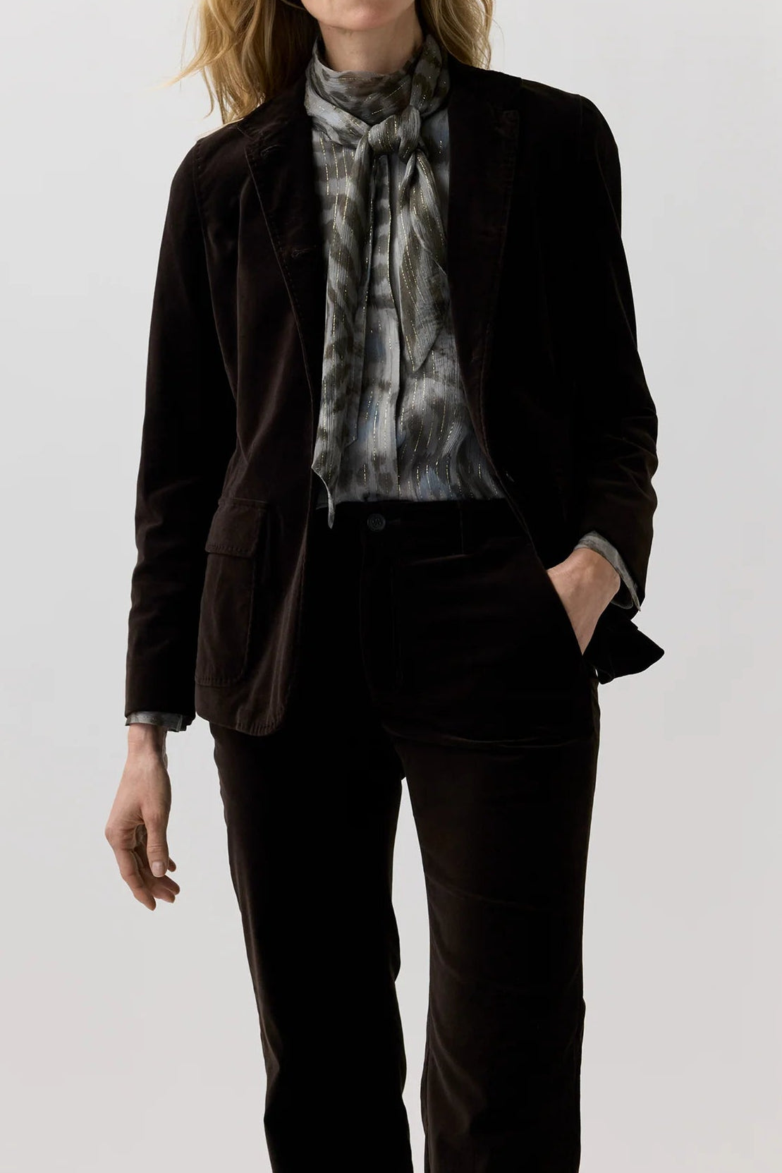 A woman modeling a dark brown velvet blazer and matching trousers, styled with a gray and white patterned silk scarf, against a plain light gray studio background.