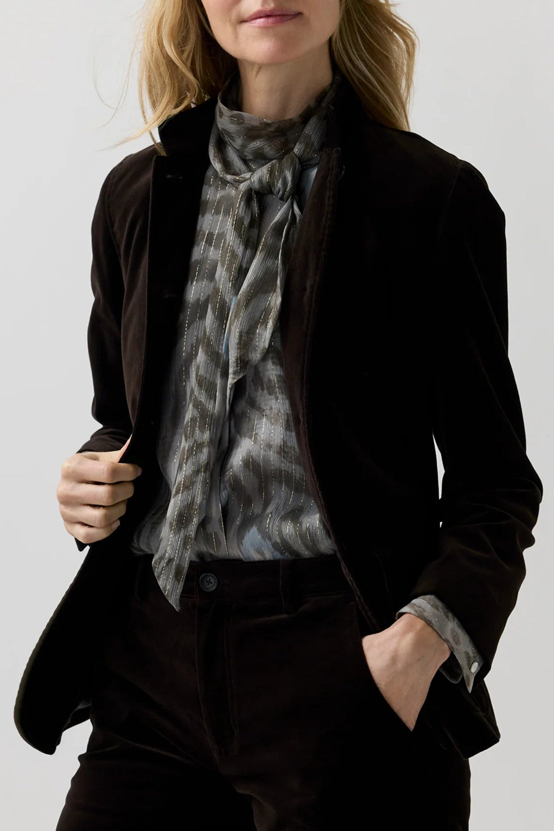 A woman modeling a dark brown velvet blazer and matching trousers, styled with a gray patterned scarf and a white collared shirt, against a light gray studio background.