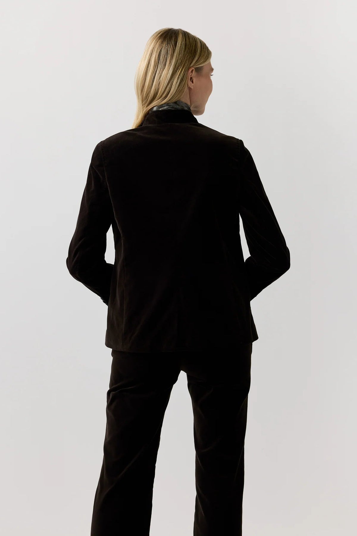 Rear view of a model wearing a black velvet blazer and matching black trousers against a light gray studio background.