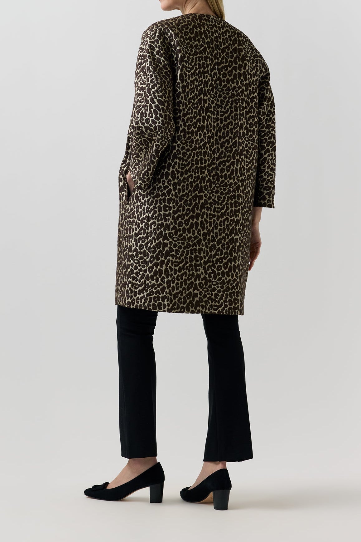 A woman shown from the back wearing a leopard-print coat over black pants and black heels, photographed against a plain light gray studio background.
