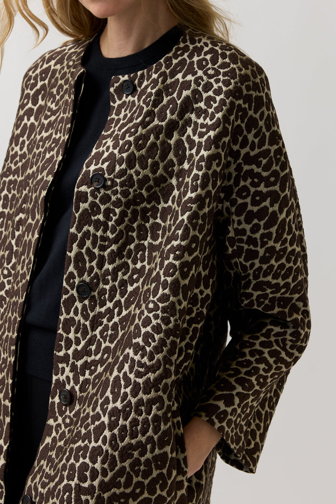 A woman wearing a brown and beige leopard-print coat over a black top, photographed against a plain light gray studio background.