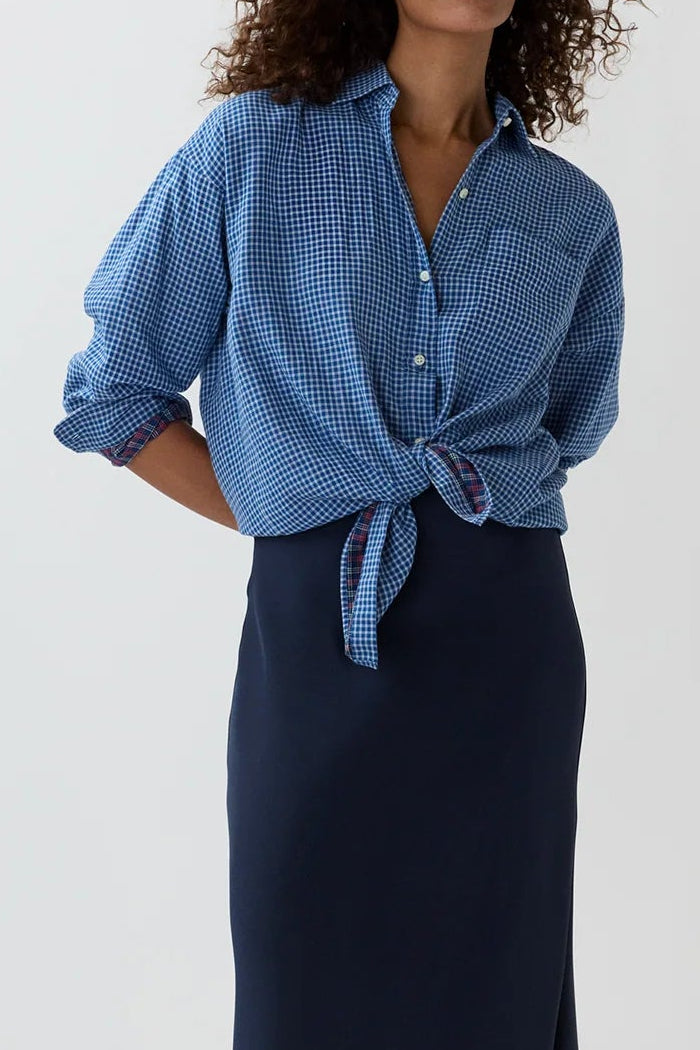 A woman modeling a blue patterned button-up shirt tied at the waist and a dark navy midi skirt, standing against a plain light gray studio background.