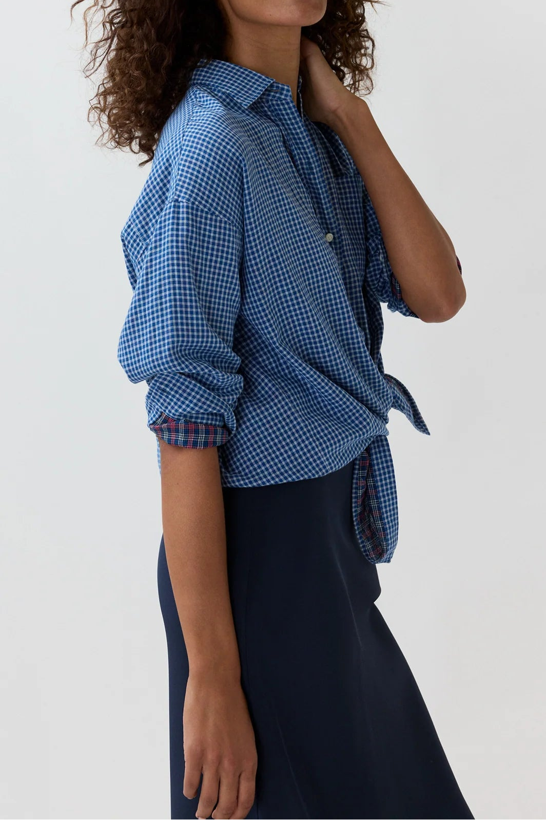 A woman with curly brown hair models a blue and white gingham button-down shirt tied at the waist, paired with a dark navy skirt, against a plain light gray studio background.