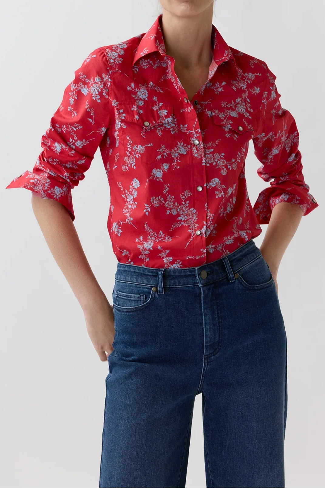 A woman modeling a red long-sleeve button-up shirt with a light blue and white floral pattern, paired with dark blue jeans, against a plain light gray background.