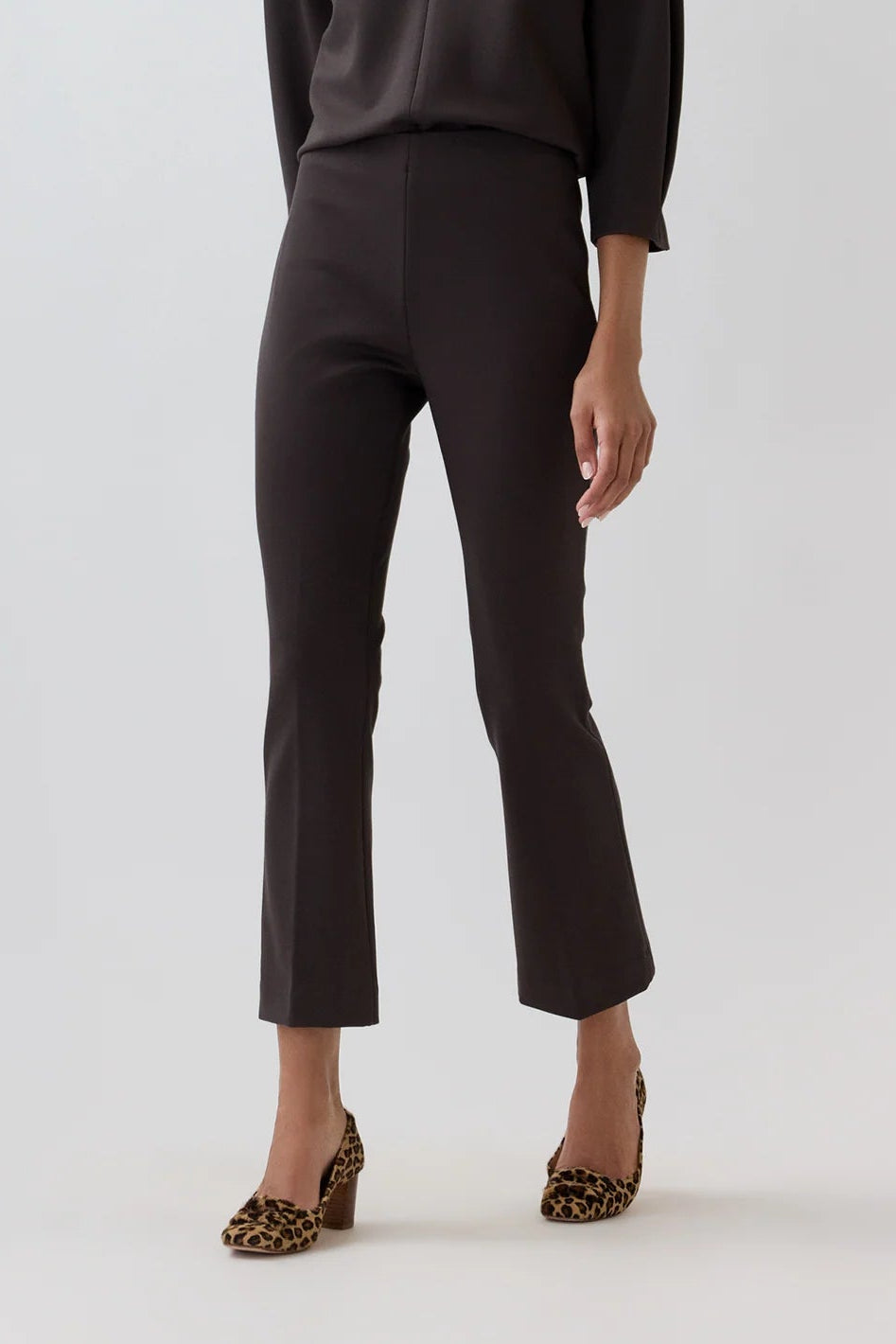 A woman modeling a dark brown long-sleeve collared shirt and matching dark brown cropped trousers, paired with leopard-print heels, against a light gray studio background.