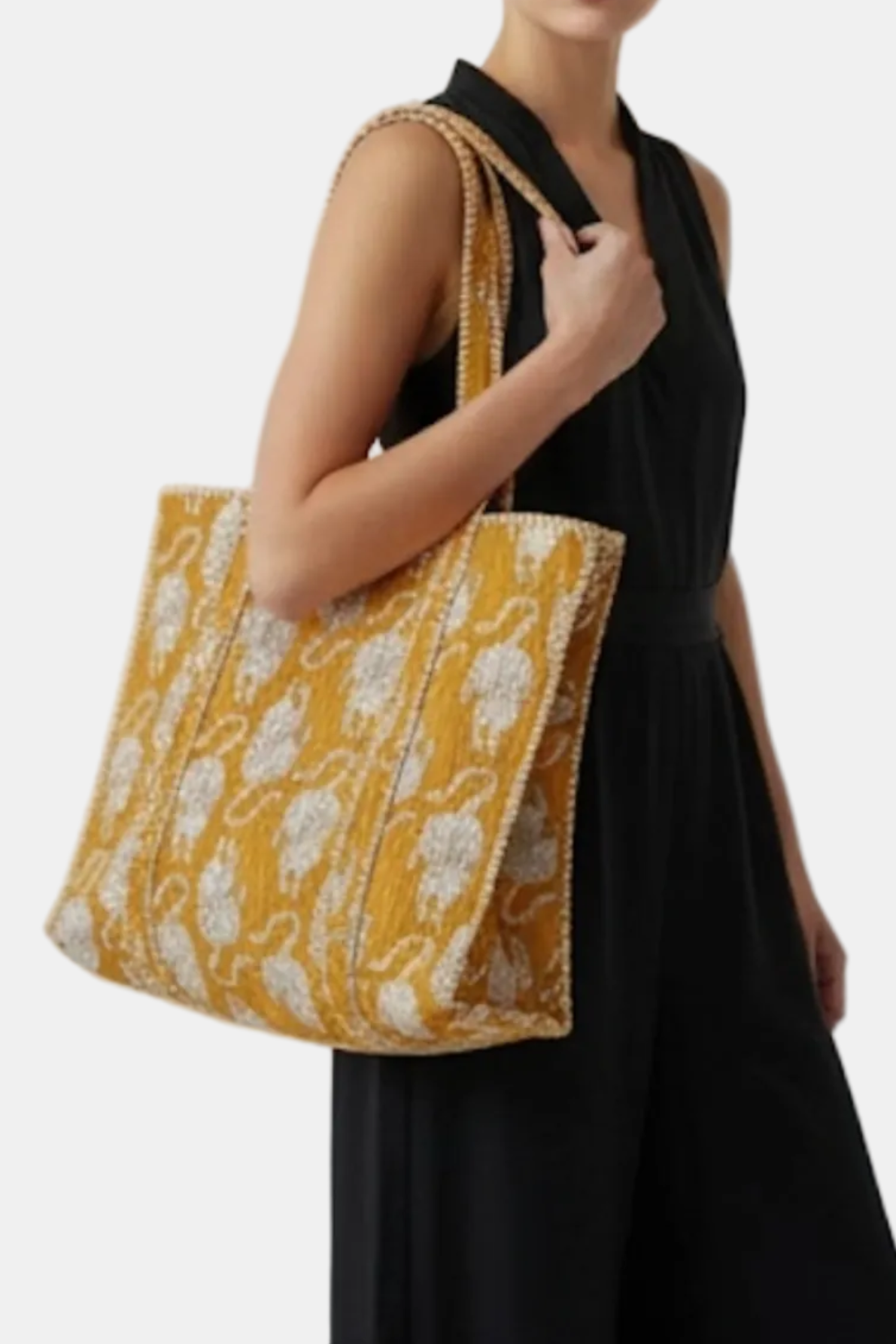 A woman wearing a black sleeveless dress carries a large mustard yellow and white patterned tote bag with gold-toned chain handles against a plain light gray background.