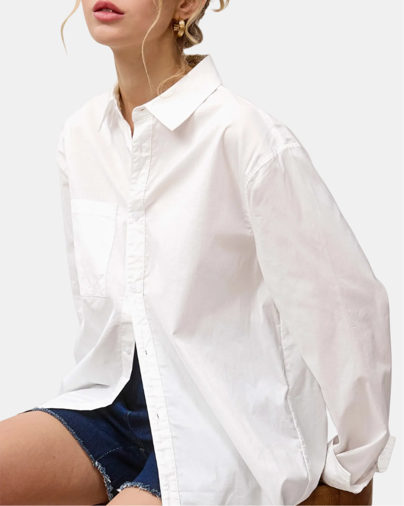 A woman wearing an oversized white long-sleeve button-up shirt with a chest pocket, paired with dark denim shorts, photographed against a plain white background.