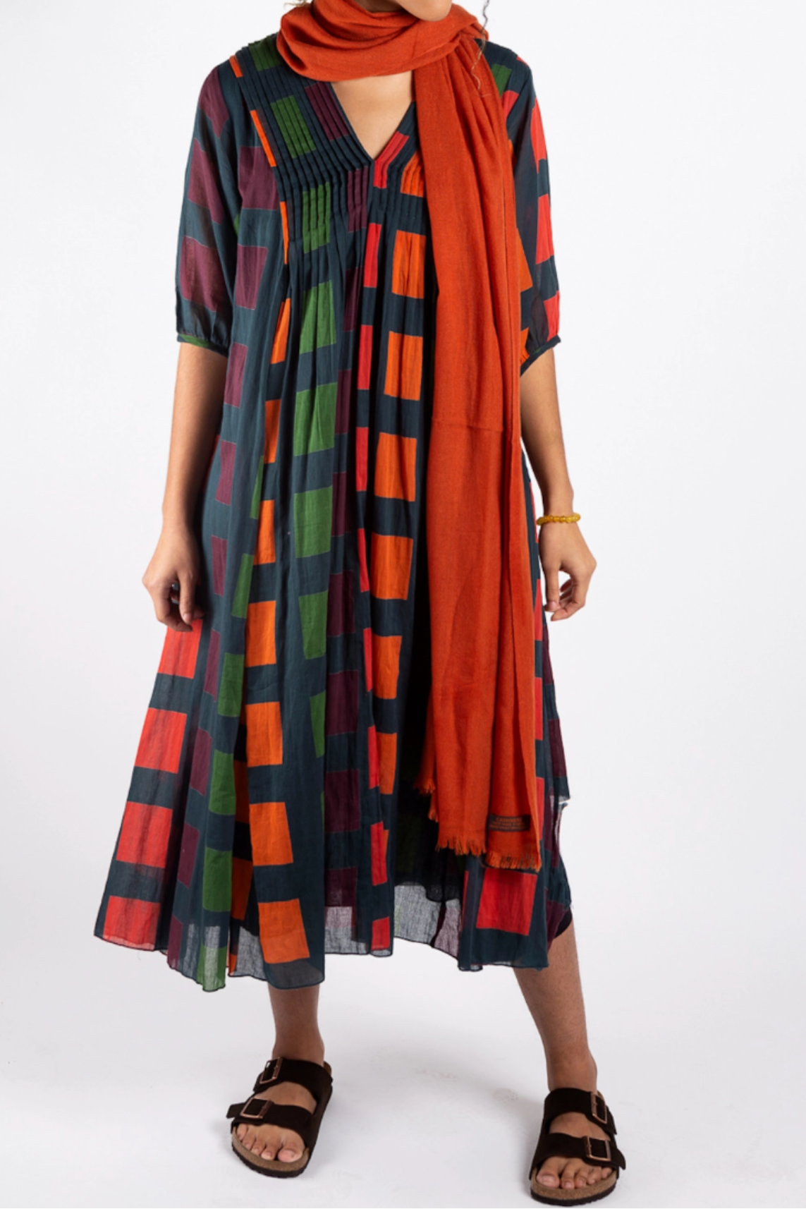A woman modeling a loose, short-sleeved midi dress with a multicolor plaid pattern in red, green, orange, and black, paired with a solid orange scarf and black sandals, against a plain white studio background.