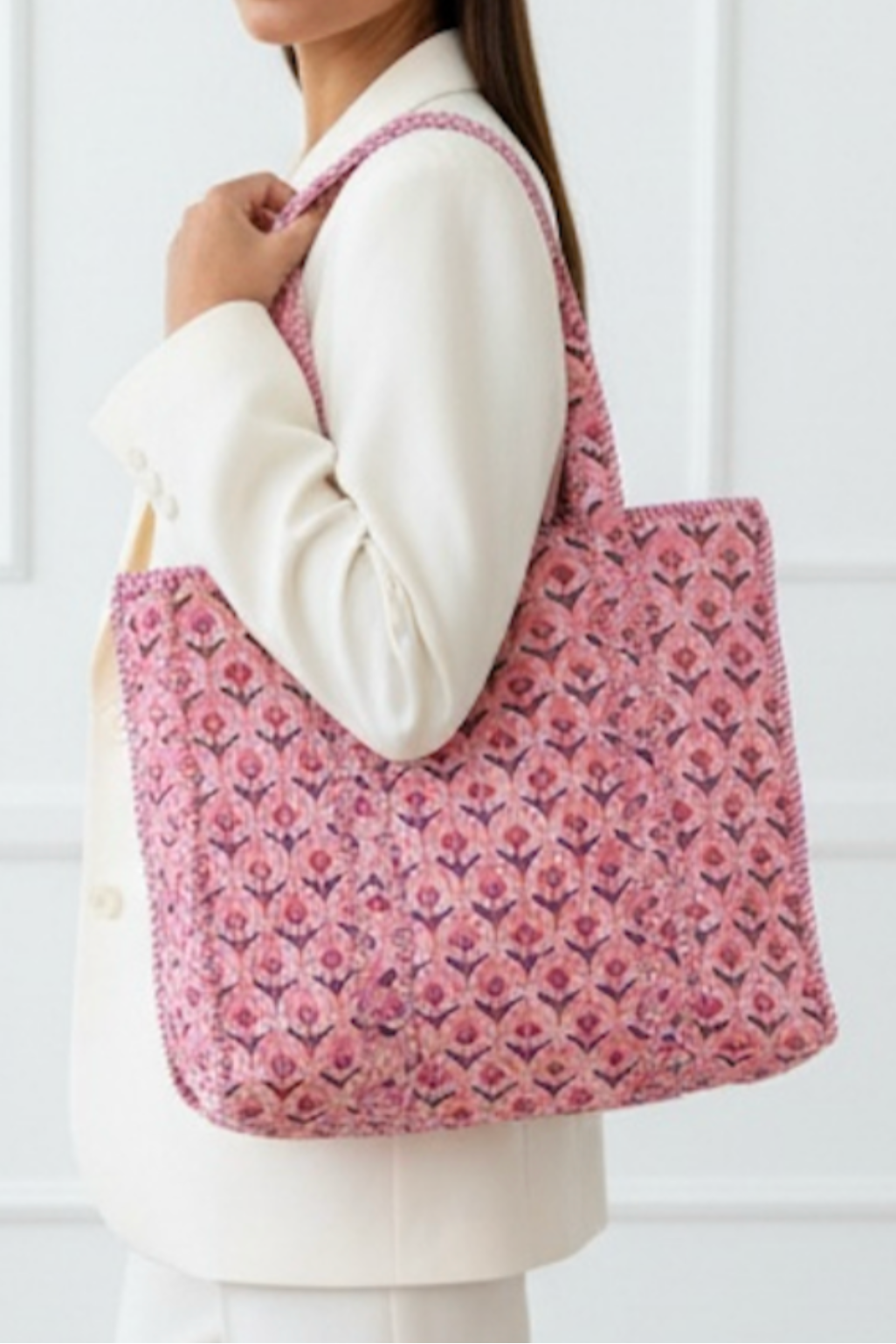 A woman wearing a white blazer and white pants carries a pink patterned woven tote bag over her shoulder, set against a white paneled wall.
