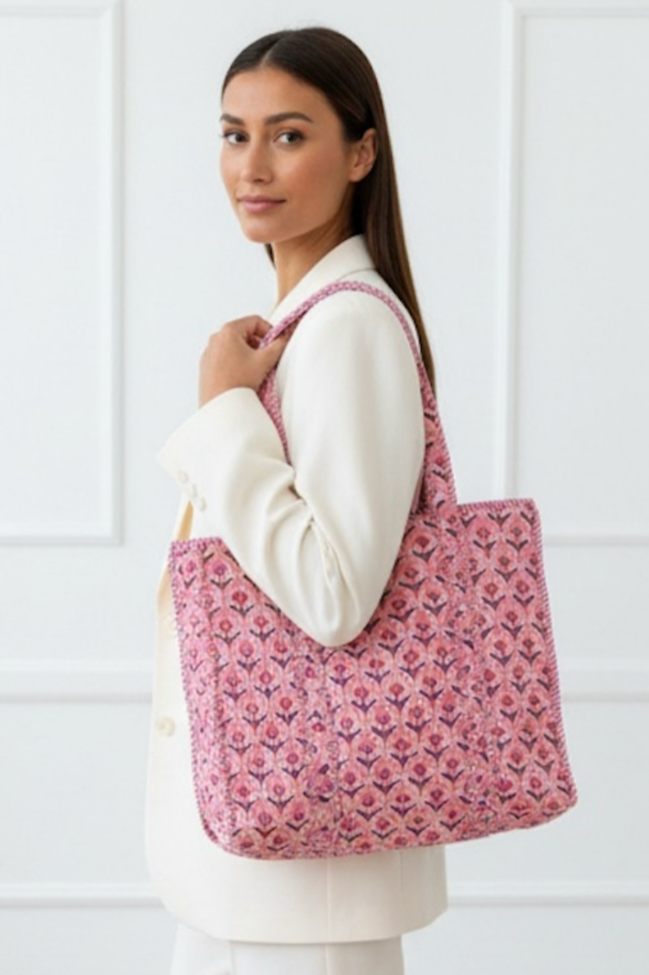 A woman wearing a white blazer and white pants carries a pink patterned woven tote bag over her shoulder, set against a white paneled wall.