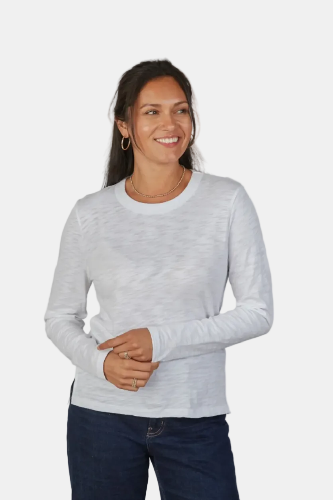 A woman wearing a light gray long-sleeve crewneck knit top and dark blue jeans, standing against a plain light gray studio background.