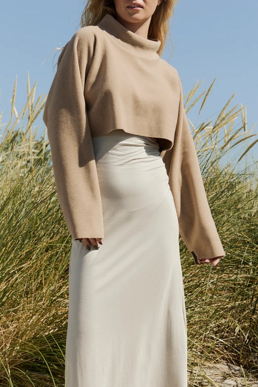 Woman standing on a sandy beach with tall grasses, wearing a beige sweater and white skirt.