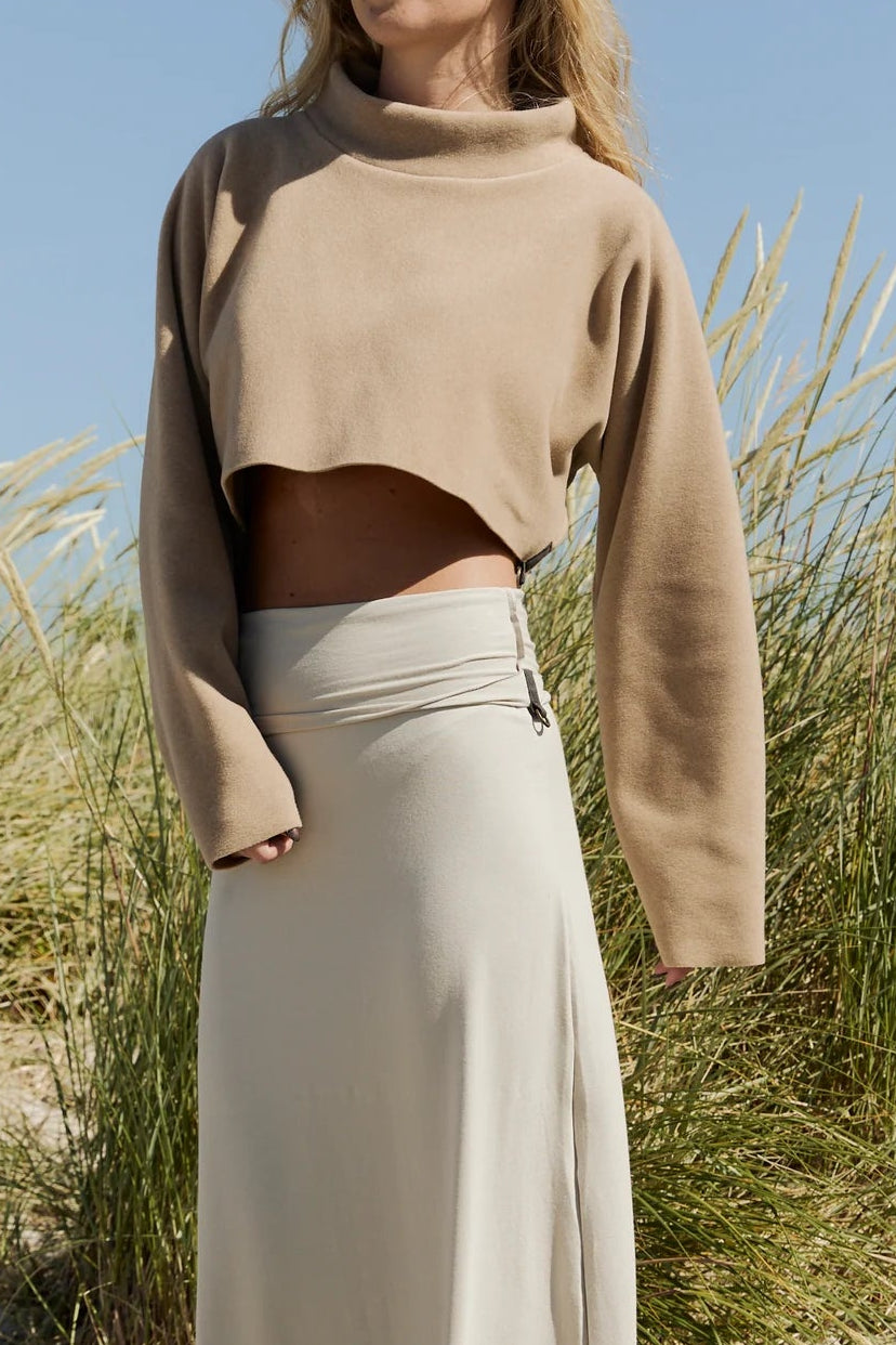 Woman in beige sweater and long skirt standing in a natural setting with tall grass and blue sky.