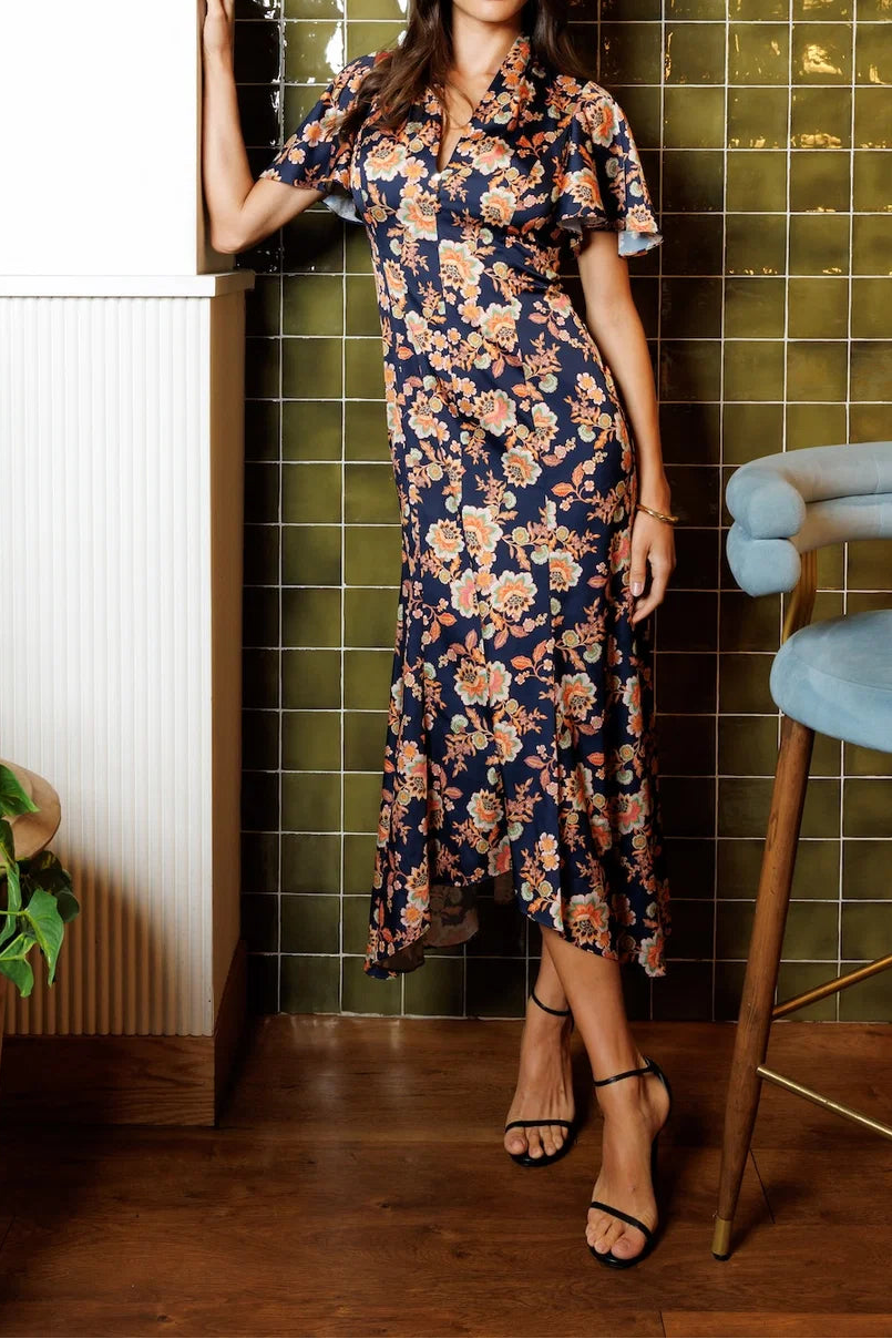 A woman models a navy blue midi dress with a beige and pink floral pattern, standing in a kitchen setting with olive green tiled walls, wooden floors, and a light blue stool.