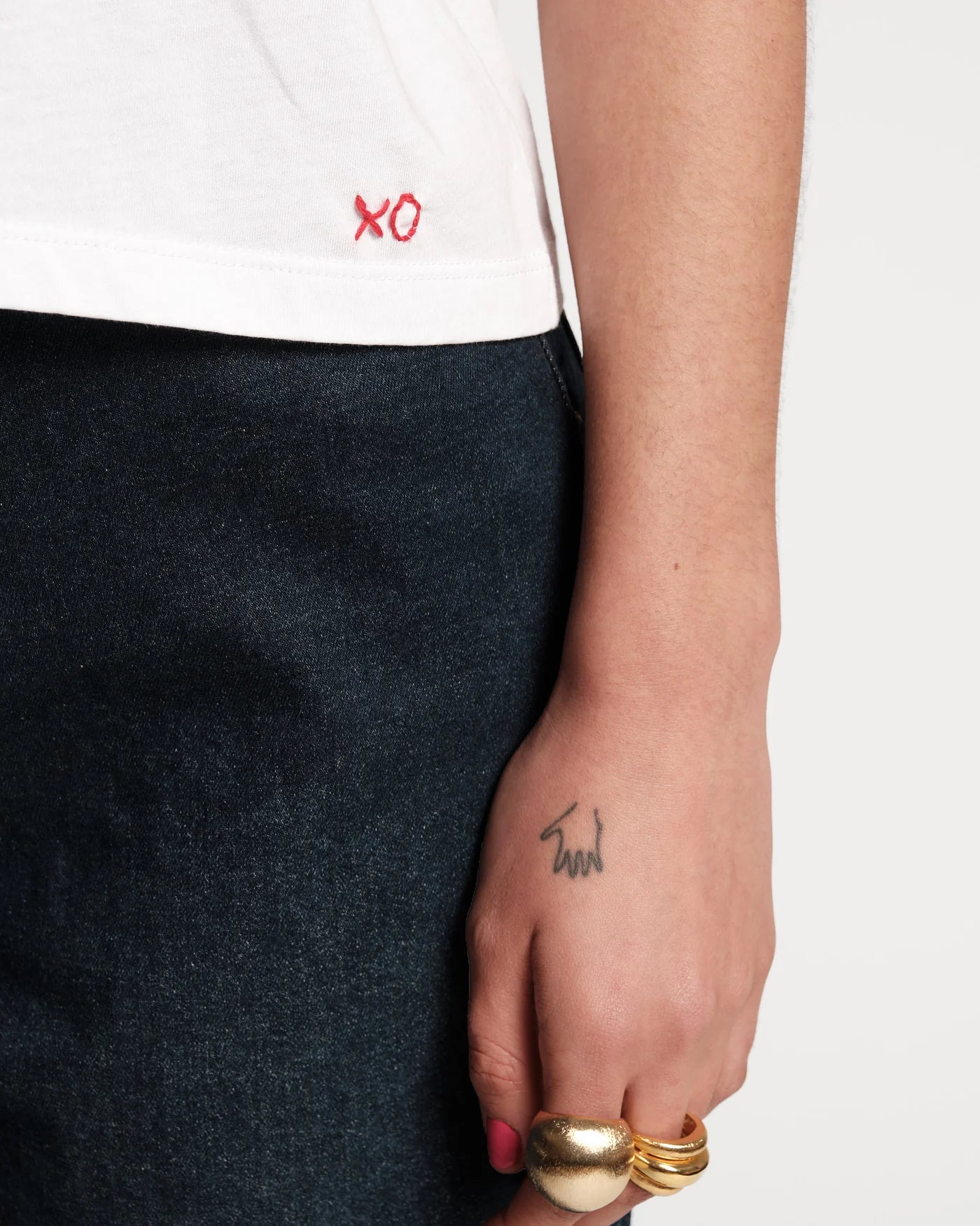 Person wearing a white shirt with red text, dark pants, and gold rings on a neutral background