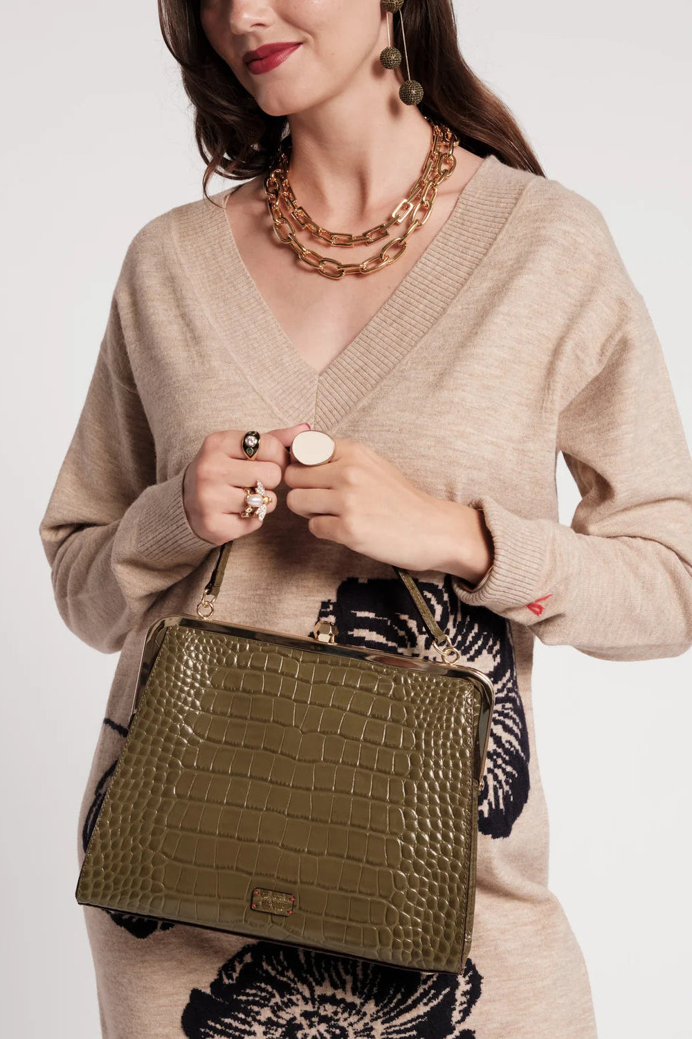 A woman wearing a beige knit sweater and holding an olive green crocodile-embossed handbag, styled with chunky gold chain necklaces and rings against a light gray studio background.