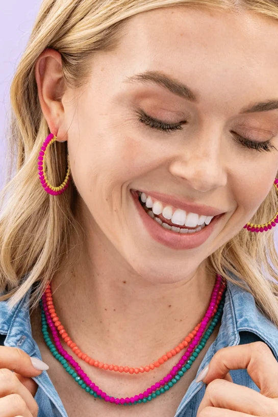 A smiling blonde woman wearing colorful beaded jewelry, including pink and orange hoop earrings and layered beaded necklaces, against a light purple background.