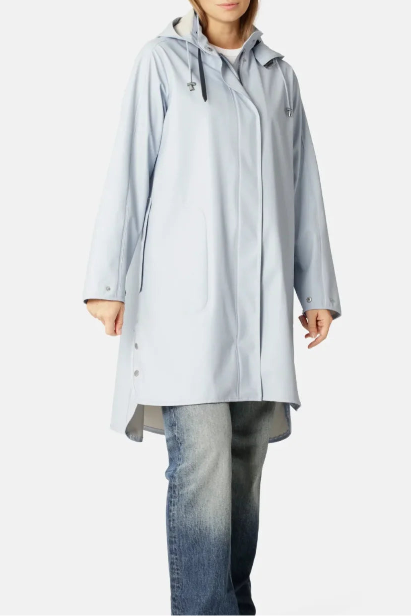 A woman modeling a light blue hooded raincoat over a white top and blue jeans, photographed against a plain light gray studio background.