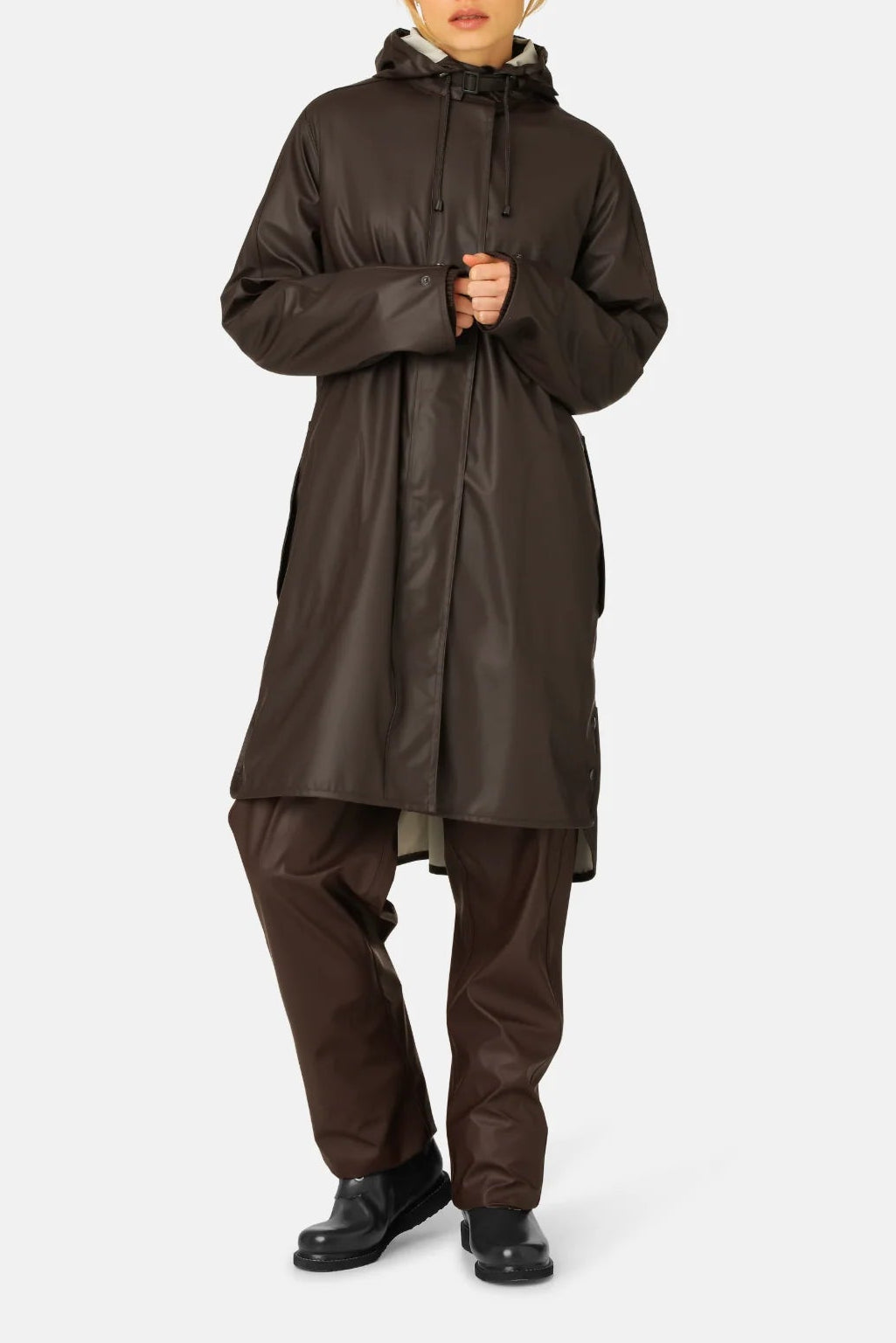 A woman modeling a long, dark brown hooded raincoat with matching dark brown pants and black shoes, standing against a light gray studio background.