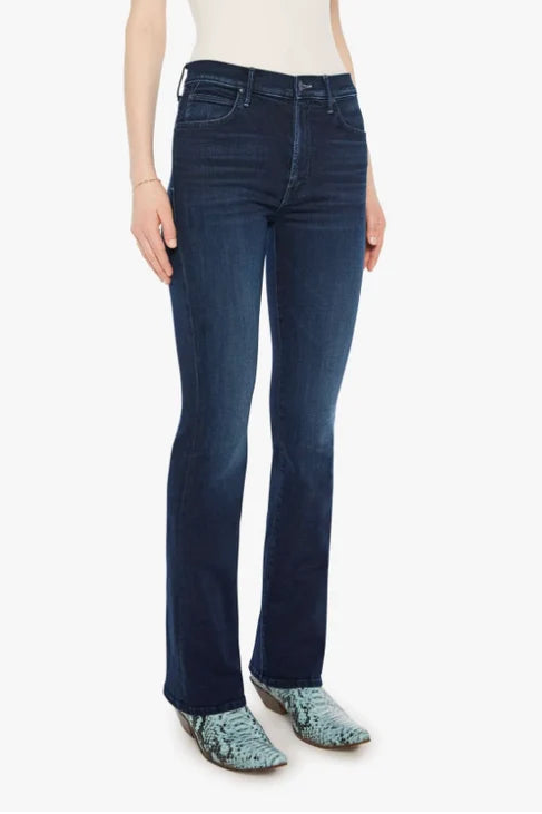 A woman modeling dark blue flare jeans with a cream ribbed tank top and patterned green shoes, standing against a plain white background.