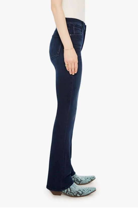 A woman shown in side profile wearing dark blue flared jeans, a cream sleeveless top, and patterned shoes against a plain light gray studio background.