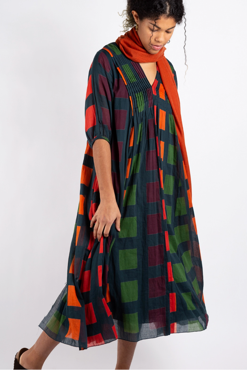 A woman modeling a loose, knee-length dress with a large geometric plaid pattern in green, orange, red, and dark navy, paired with a matching orange scarf, against a plain white studio background.
