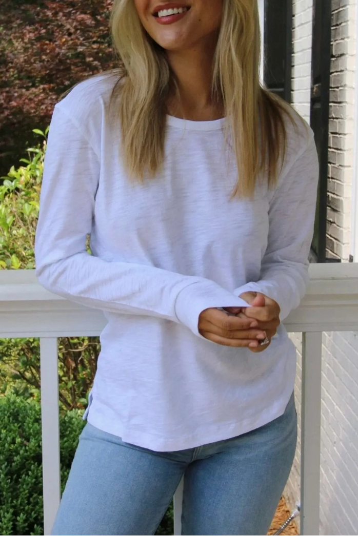 A woman with blonde hair smiling while wearing a white long-sleeve crewneck shirt and light blue jeans, standing on a white porch with trees and a house in the background.