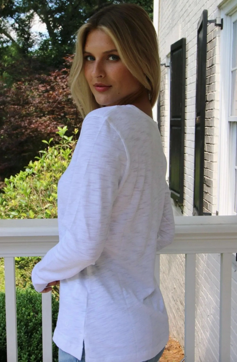 A woman with blonde hair is shown from the back wearing a white long-sleeve linen-blend top on a white porch with a brick house and greenery in the background.
