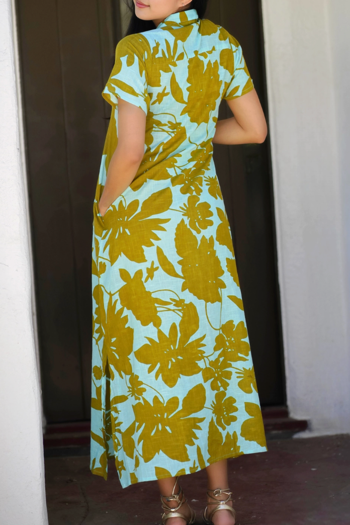 A woman wearing a light blue and mustard yellow floral print cheongsam dress, standing in front of a doorway.