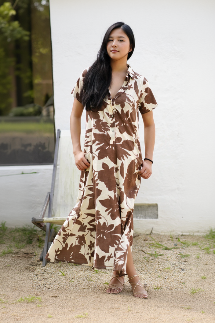A woman wearing a short-sleeve button-front maxi dress with a large brown floral print on a cream background, standing outdoors in front of a white wall.