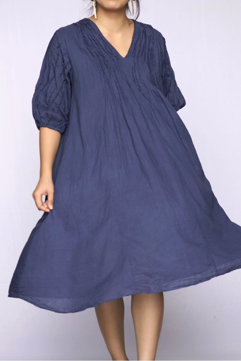 A woman wearing a loose, flowy midi dress in a dusty blue-purple color, standing against a plain light background.