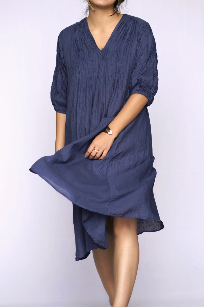 A woman wearing a loose, short-sleeved navy blue dress with a V-neckline and gathered sleeves, standing against a plain light background.