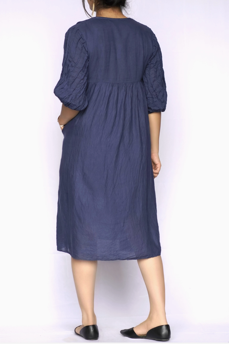 A woman viewed from the back wearing a dark blue midi dress with three-quarter puffed sleeves and black flat shoes, set against a plain white studio background.