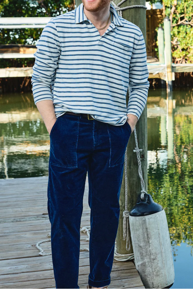A man standing on a wooden dock by the water wearing a navy and white striped long-sleeve polo shirt, dark blue corduroy trousers, and a navy baseball cap, with a white fender hanging beside him.