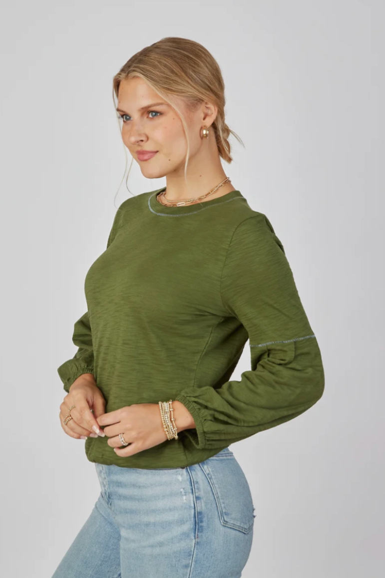 A woman wearing an olive green long-sleeve top and light blue jeans, standing against a plain light gray studio background.