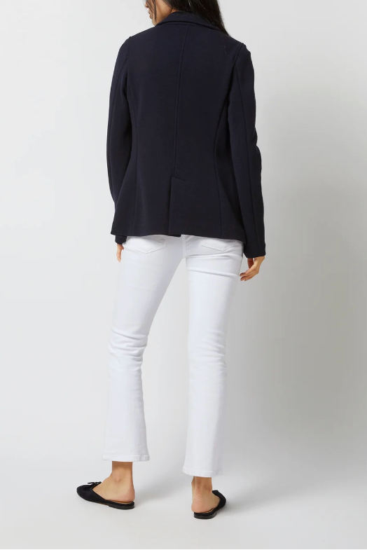 A woman shown from the back wearing a dark navy blazer, white cropped pants, and black flat shoes against a light gray studio background.