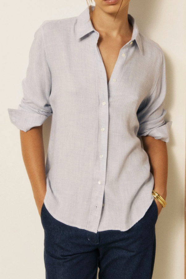 A woman wearing a light gray linen button-down shirt with rolled-up sleeves and dark blue jeans, standing against a split cream and wood-paneled wall.
