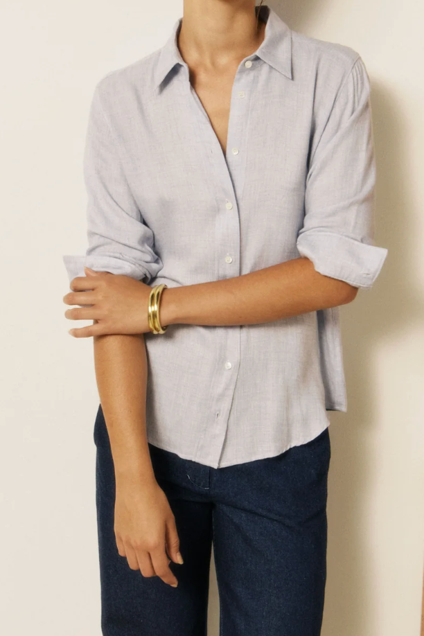 A woman wearing a light blue and white micro-check button-down shirt with dark blue jeans, standing against a neutral off-white wall with a wooden panel on the right.