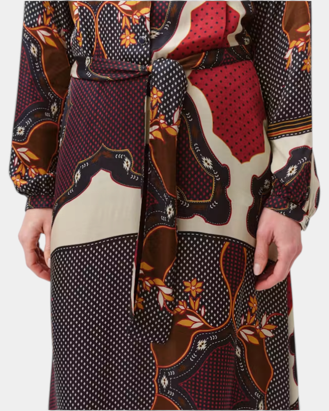 A close-up of a person wearing a long-sleeve belted wrap dress with a complex multicolor patchwork print featuring red, brown, black, and cream sections.