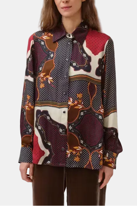 A woman modeling a long-sleeve button-up shirt with an abstract, colorful print featuring red, brown, navy, and cream shapes, paired with dark brown pants against a light gray studio background.