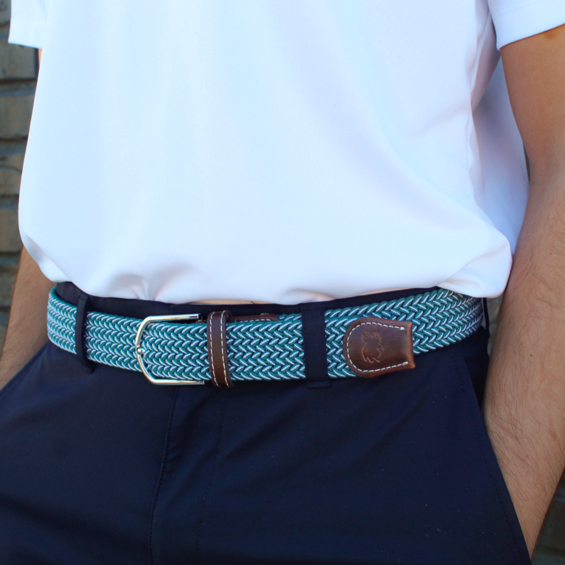 A close-up of a person wearing a woven belt with a teal and white herringbone pattern, brown leather accents, and a silver buckle, paired with a white polo shirt and navy pants.