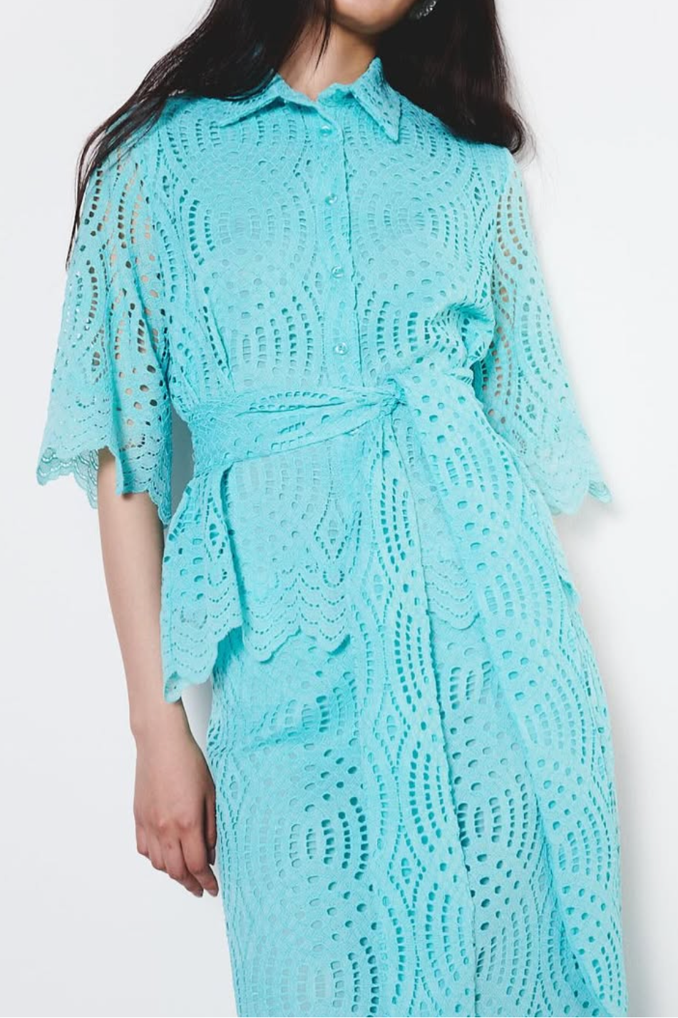 A woman modeling a light turquoise blue crochet lace shirt dress with a collar, short sleeves, and a matching tie belt, set against a plain white studio background.