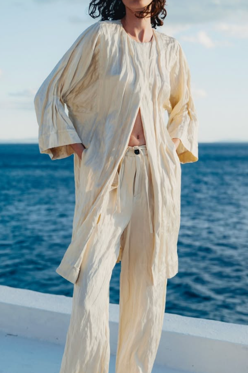 A model wearing a cream-colored crinkled linen two-piece set consisting of a long-sleeve button-up shirt and matching wide-leg trousers, standing on a white balcony with the blue sea and sky in the background.
