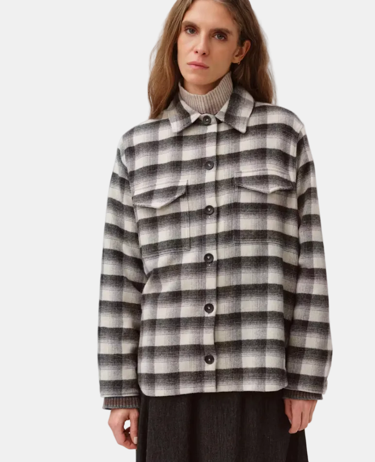 A woman wearing a black and off-white checkered button-up shirt jacket over a ribbed beige turtleneck, photographed against a plain light gray studio background.
