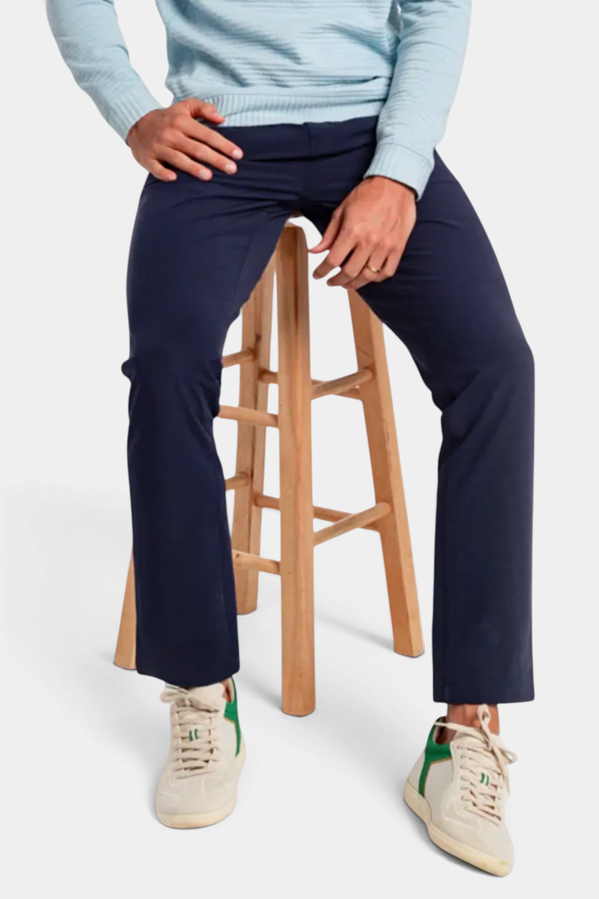 Person wearing navy pants and a light blue sweater sitting on a wooden stool.