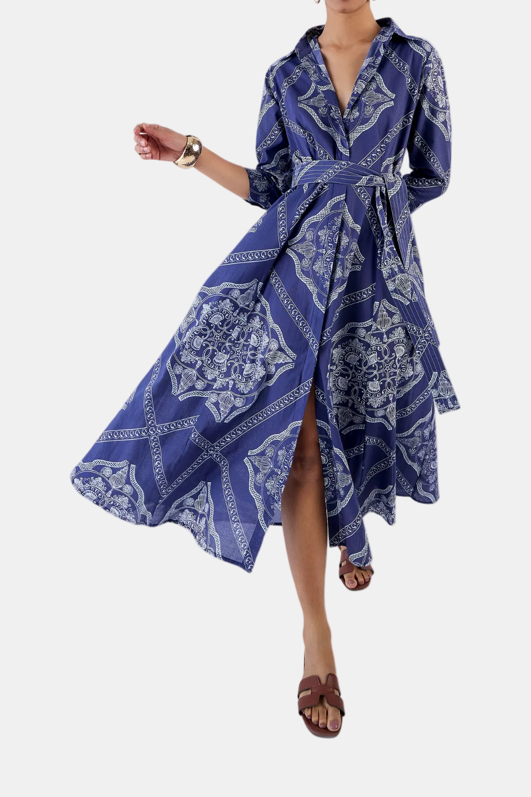 A woman modeling a blue and white patterned midi dress with a tie waist and long sleeves, paired with brown strappy sandals, against a light gray background.