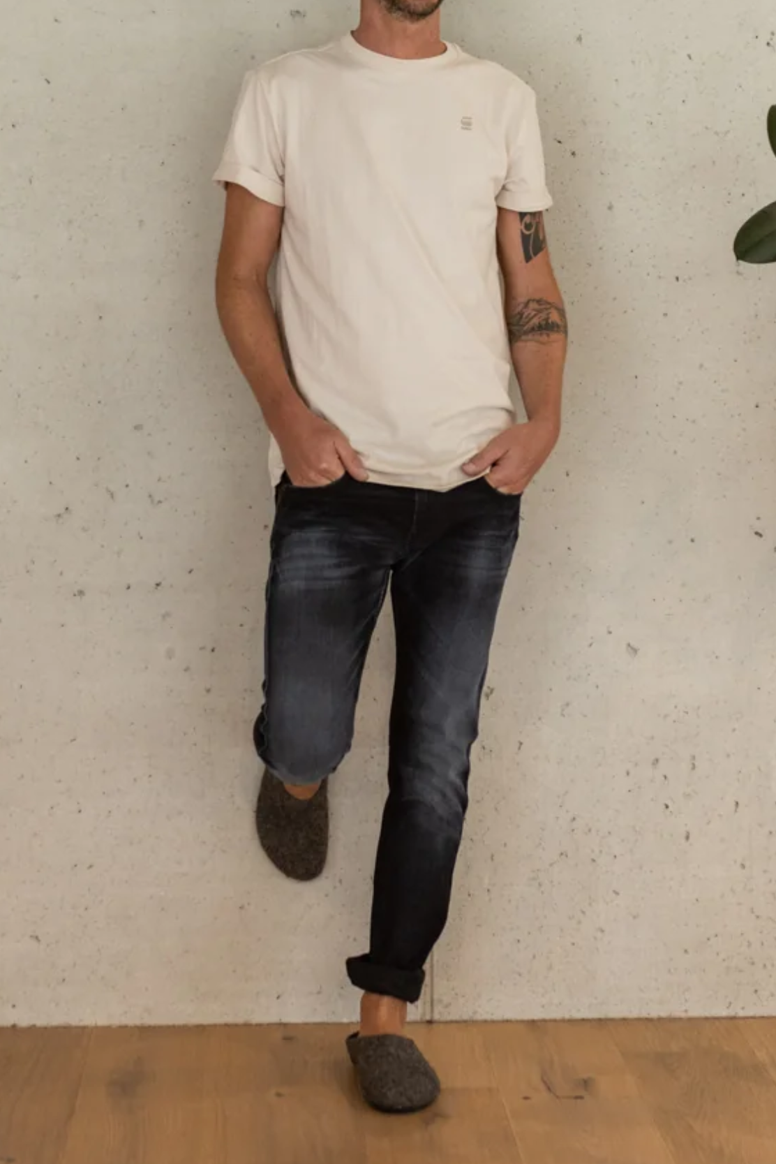 Man wearing a white t-shirt and dark jeans against a textured wall.