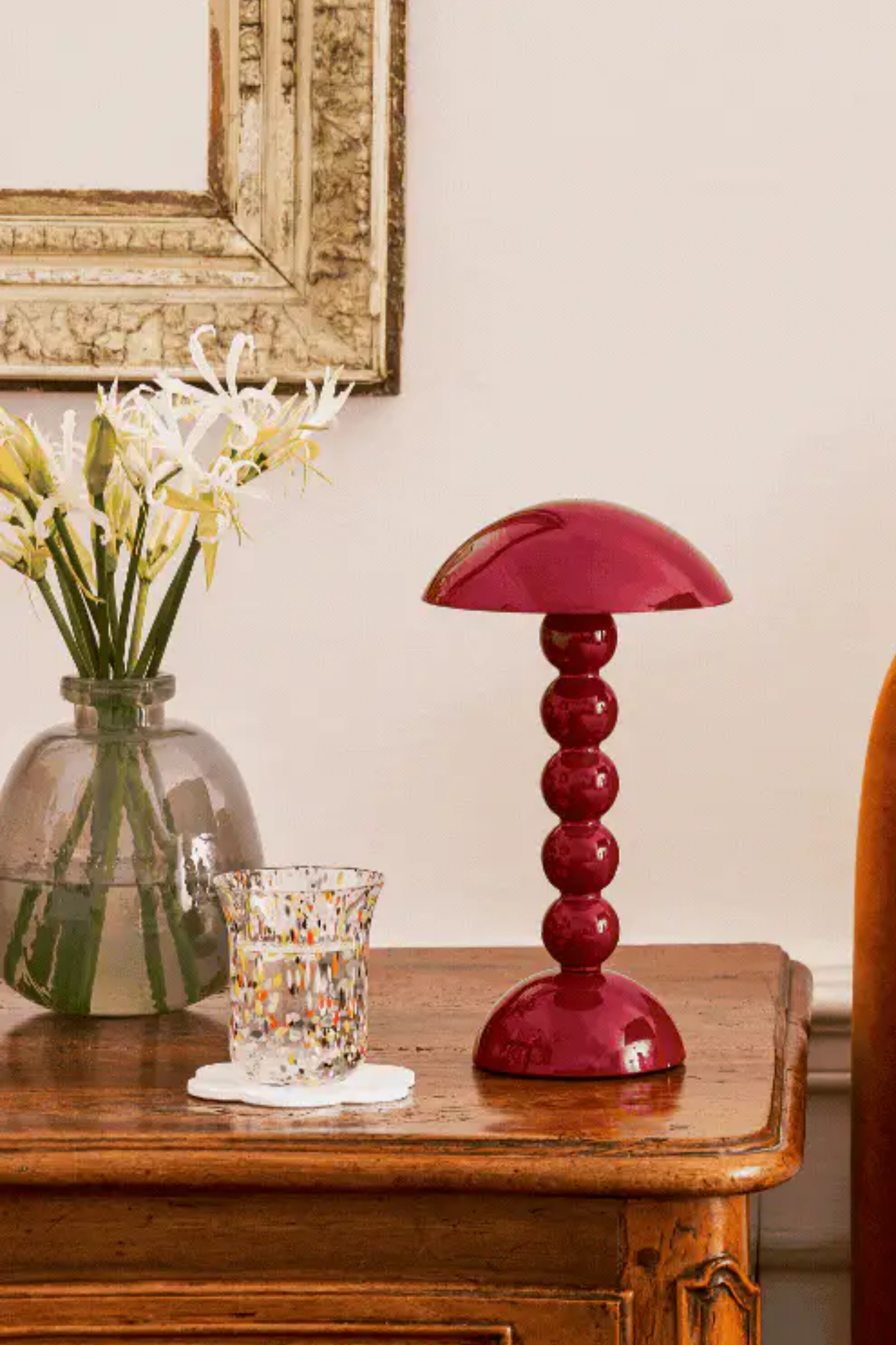 Red table lamp on a wooden surface with a vase of flowers and a candle in the background.