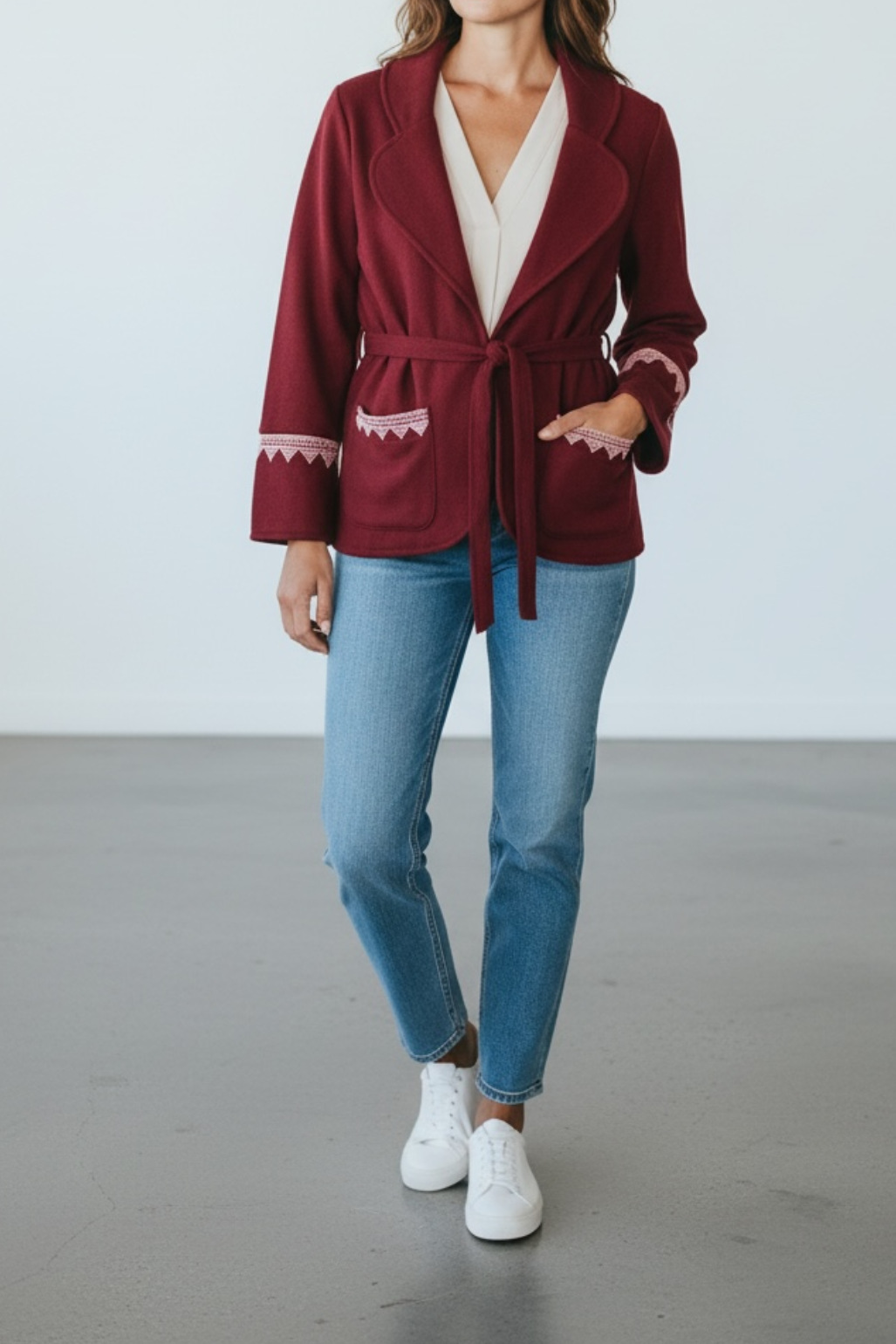 Woman wearing a burgundy robe with white trim, light blue jeans, and white sneakers on a plain background