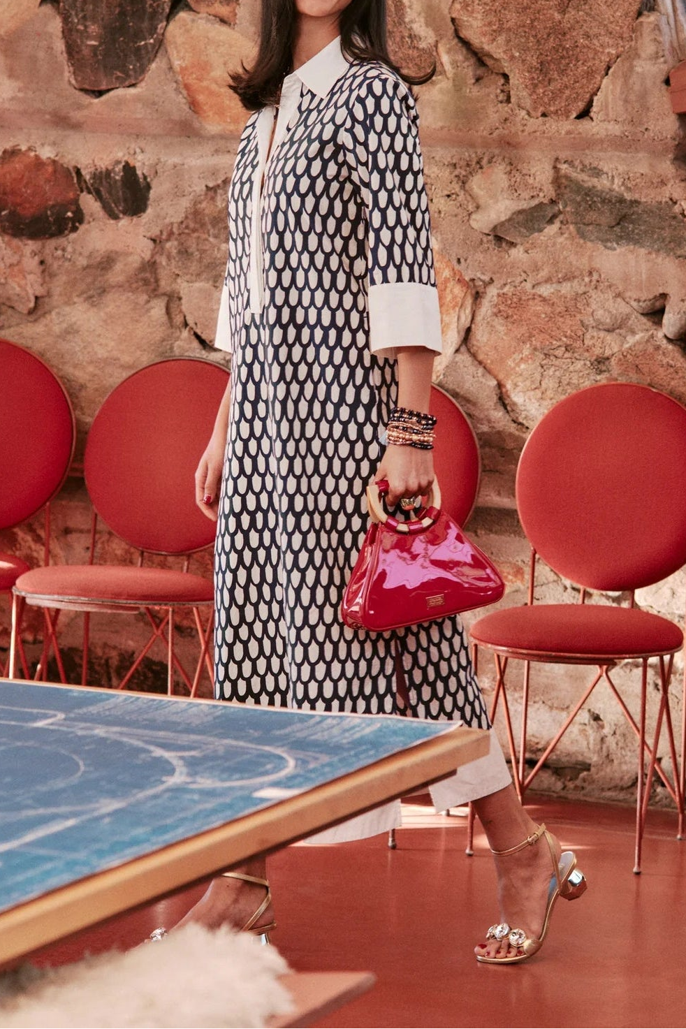 Woman in a patterned dress holding a red handbag in a room with red chairs and a stone wall.